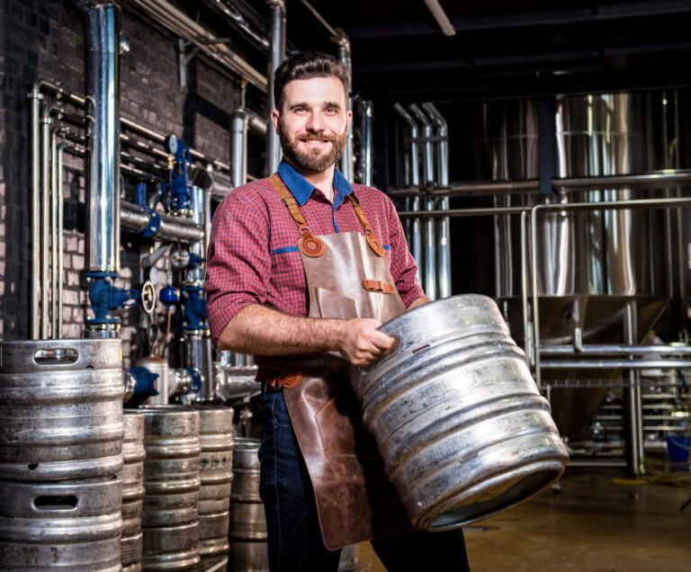 Macho joven cervecero en delantal de cuero tiene barril con cerveza artesanal en la fábrica de cerveza moderna